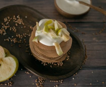 From above of tasty homemade pancakes made of buckwheat topped with sour cream and apples served on wooden table in kitchen