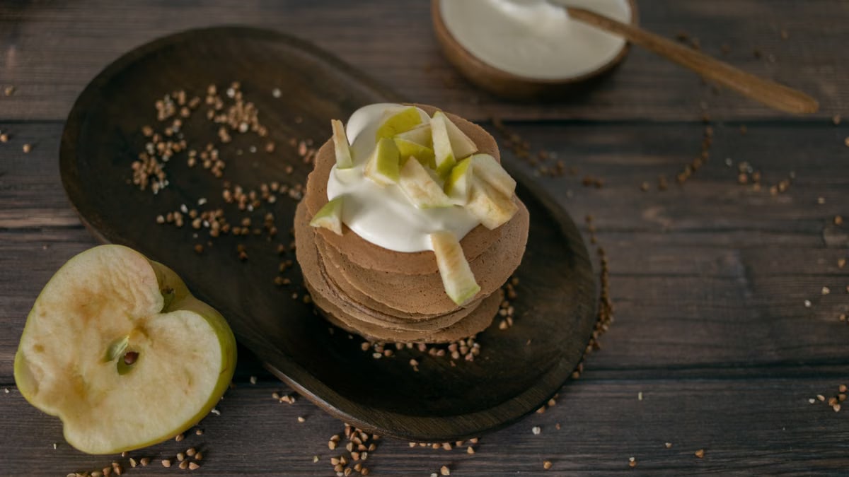 From above of tasty homemade pancakes made of buckwheat topped with sour cream and apples served on wooden table in kitchen